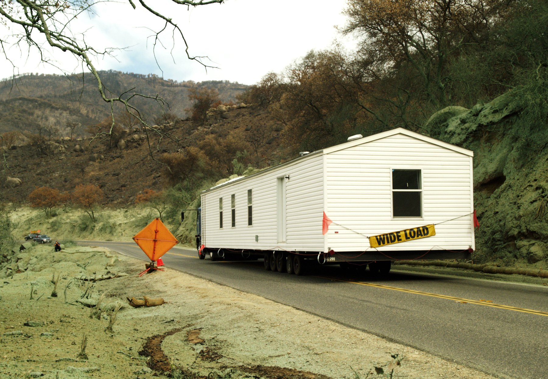 A single-wide manufactured home on the road under a wide load banner, escort flag ahead